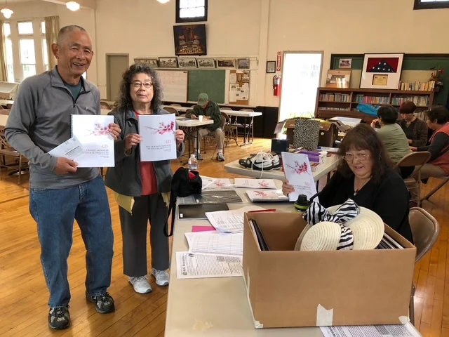 Seven people gather in the HJCI community hall as attendees display copies of the 2017 70th Anniversary book.